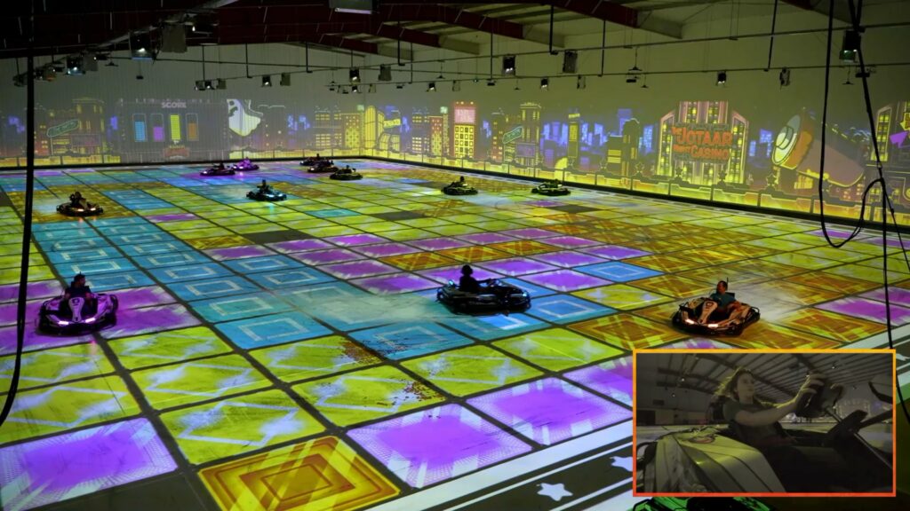Karts racing on a coloured grid projected across the arena floor, with an onboard camera inset