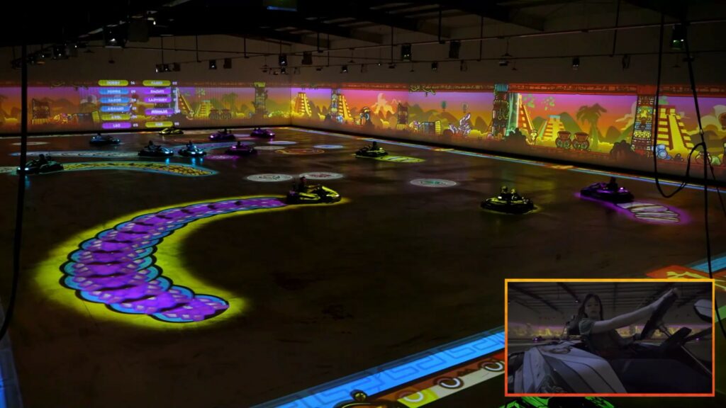 Karts following a purple curve projected on the arena floor, with an onboard camera inset